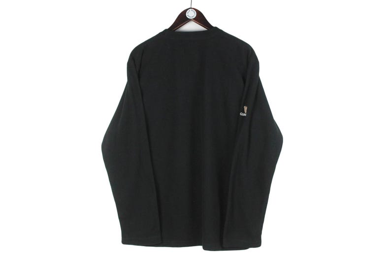 Op de afbeelding: Zwart shirt met lange mouwen hangend aan een houten kleerhanger. Het shirt heeft een klein logo op de mouw. Het shirt is gemaakt van een zacht materiaal.