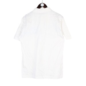 May include: A white short-sleeved polo shirt hanging on a wooden hanger. The shirt is plain and appears to be made of a lightweight fabric. The back of the shirt is visible, showing the collar and the upper back.