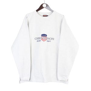 Puede incluir: Una sudadera gris claro con mangas largas, colgada de una percha de madera. La parte delantera presenta un logotipo con el texto "CHENE NON SPORT DEPT" en un diseño de escudo con detalles en rojo, blanco y azul. La sudadera es de material suave.