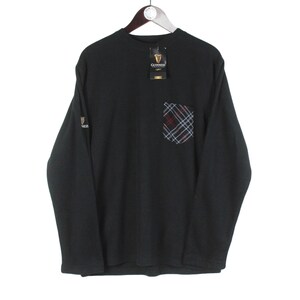 Op de afbeelding: Zwart shirt met lange mouwen, een ruitvormige zak en een Guinness-logotag. Het shirt heeft een Guinness-logo op de linkermouw. De zak heeft een ruitpatroon in rood, blauw en wit. Het shirt hangt aan een houten hanger.