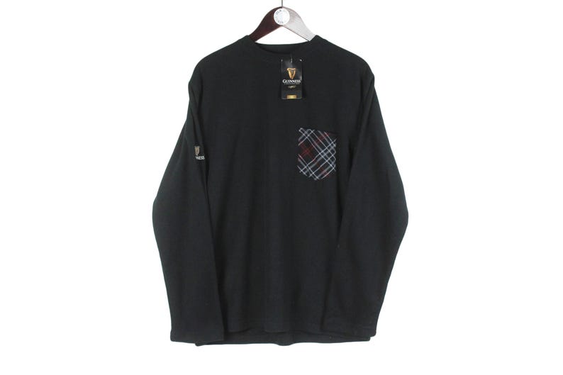Op de afbeelding: Zwart shirt met lange mouwen, een ruitvormige zak en een Guinness-logotag. Het shirt heeft een Guinness-logo op de linkermouw. De zak heeft een ruitpatroon in rood, blauw en wit. Het shirt hangt aan een houten hanger.