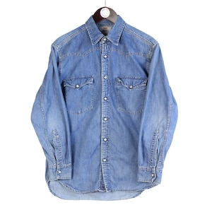 Puede incluir: Camisa vaquera azul claro con mangas largas, cierre de botones y dos bolsillos en el pecho. La camisa está colgada de una percha de madera. El tejido vaquero tiene un aspecto descolorido.