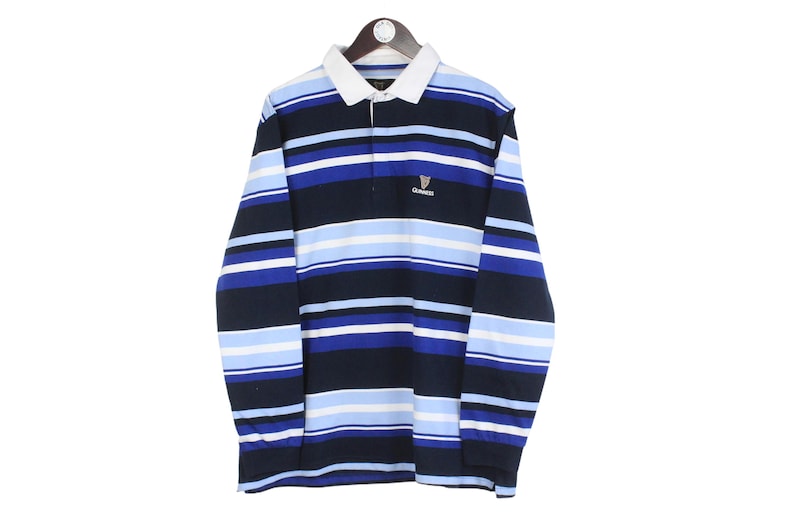 May include: A striped rugby shirt in navy, light blue, and white, with a white collar and a Guinness logo. The long-sleeved shirt is hanging on a wooden hanger. The shirt's design features horizontal stripes.