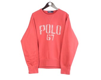 May include: Coral-coloured sweatshirt with the word "POLO" and the number "67" printed in white. The crew neck sweatshirt is hanging on a black hanger. The garment has ribbed cuffs and hem.