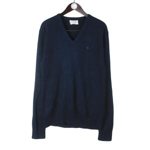 Può includere: Un maglione blu navy con scollo a V e maniche lunghe, appeso a una gruccia di legno. Il maglione ha un piccolo stemma blu sul petto. L'etichetta è visibile allo scollo. Il maglione è realizzato in materiale morbido.