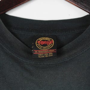 May include: Black t-shirt with a crew neck and a tag featuring the Guinness logo. The tag reads "Official Merchandise" and "Chest Size XXL 42/44 106/111cm". The shirt is hanging on a wooden hanger.