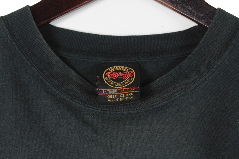May include: Black t-shirt with a crew neck and a tag featuring the Guinness logo. The tag reads "Official Merchandise" and "Chest Size XXL 42/44 106/111cm". The shirt is hanging on a wooden hanger.