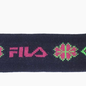 May include: Navy blue knit headband with the word "FILA" in pink. The headband features pink and green floral and arrow designs. Pink and green yarn strands extend from one end.