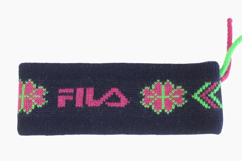 May include: Navy blue knit headband with the word "FILA" in pink. The headband features pink and green floral and arrow designs. Pink and green yarn strands extend from one end.