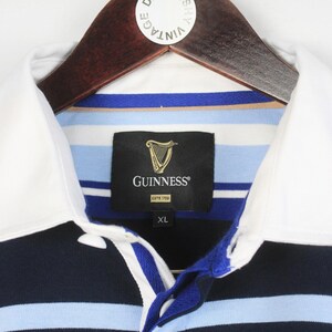 May include: A close-up of a rugby shirt with a white collar and the Guinness logo. The shirt has blue, white, and navy stripes. The label indicates an XL size.