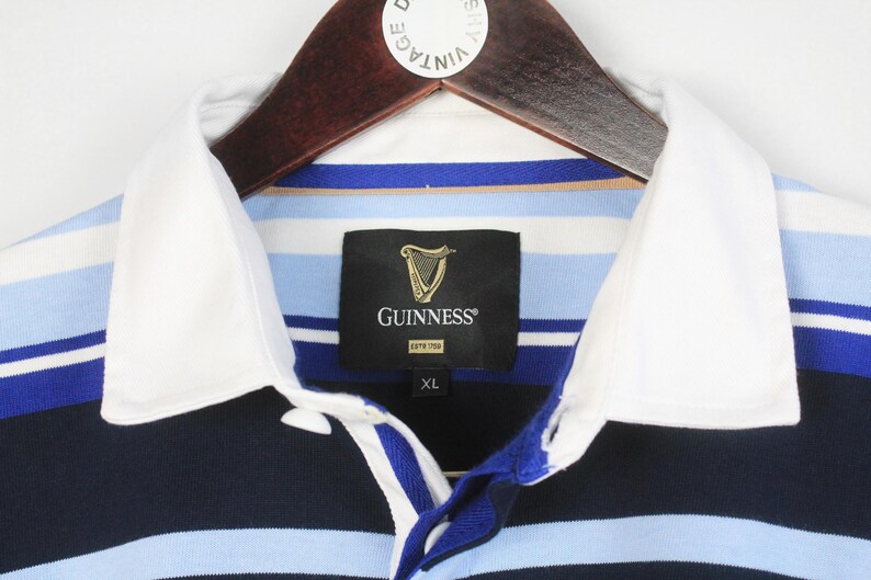 May include: A close-up of a rugby shirt with a white collar and the Guinness logo. The shirt has blue, white, and navy stripes. The label indicates an XL size.