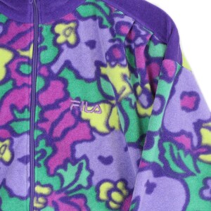 Puede incluir: Una chaqueta de forro polar con cremallera y un vibrante estampado floral en tonos morado, verde brillante y amarillo. La prenda incluye un cierre de cremallera morado y el logotipo de la marca Fila bordado en morado en el lado izquierdo del pecho.
