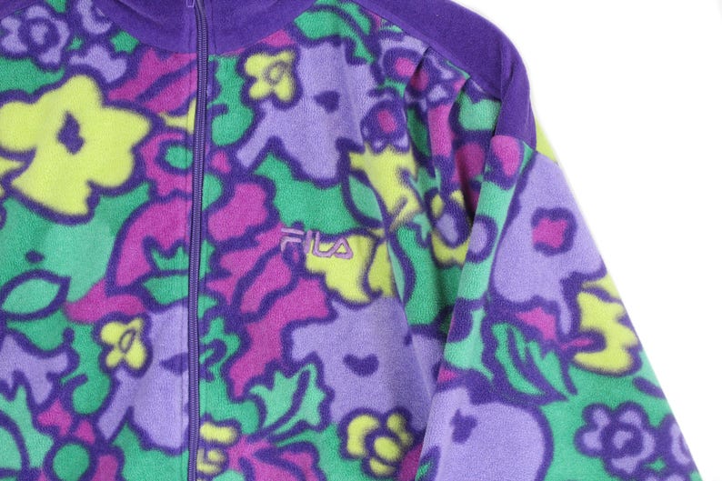Puede incluir: Una chaqueta de forro polar con cremallera y un vibrante estampado floral en tonos morado, verde brillante y amarillo. La prenda incluye un cierre de cremallera morado y el logotipo de la marca Fila bordado en morado en el lado izquierdo del pecho.