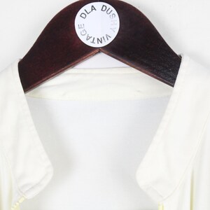 May include: A sleeveless white top with a collar, hanging on a dark brown wooden hanger. A circular white sticker is attached to the hanger featuring the black text DLA DUSZY VINTAGE arranged in a circular pattern around the center of the sticker.