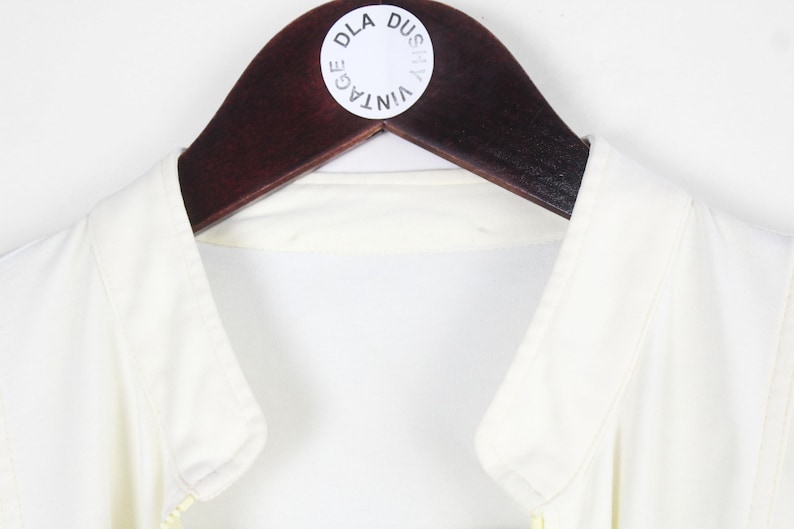 May include: A sleeveless white top with a collar, hanging on a dark brown wooden hanger. A circular white sticker is attached to the hanger featuring the black text DLA DUSZY VINTAGE arranged in a circular pattern around the center of the sticker.