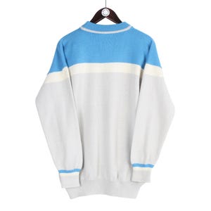Puede incluir: Un su&eacute;ter vintage con un dise&ntilde;o de bloques de color. La parte superior de los hombros es azul claro, con una franja blanca debajo, y el resto del su&eacute;ter es gris claro. Los pu&ntilde;os y el cuello tienen rayas azul claro.