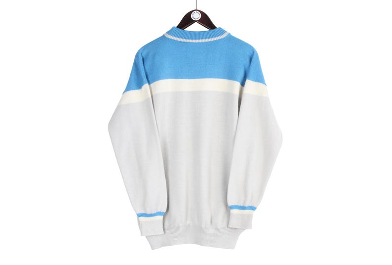 Puede incluir: Un su&eacute;ter vintage con un dise&ntilde;o de bloques de color. La parte superior de los hombros es azul claro, con una franja blanca debajo, y el resto del su&eacute;ter es gris claro. Los pu&ntilde;os y el cuello tienen rayas azul claro.