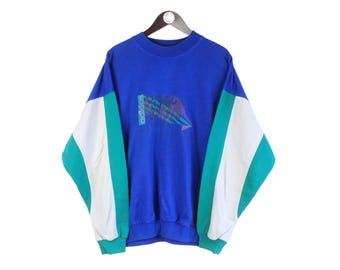 Könnte beinhalten: Ein blaues Vintage-Sweatshirt mit weiß-türkisfarbenen Farbblockärmeln. Auf der Vorderseite befindet sich ein grafisches Design mit dem Adidas-Logo. Das Sweatshirt hängt an einem Holzbügel.