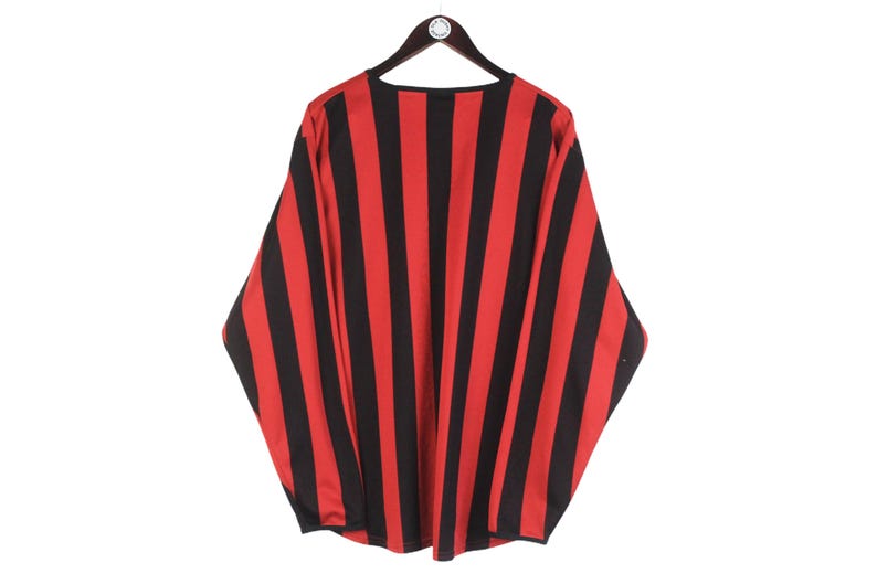 Puede incluir: Una camisa de manga larga con rayas verticales rojas y negras, colgada de una percha de madera. El patr&oacute;n de la camisa es consistente en toda la prenda, creando una declaraci&oacute;n visual audaz. La tela parece ser ligera.