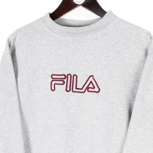 Puede incluir: Sudadera gris claro con el nombre de la marca "FILA" bordado en rojo y rojo oscuro. La sudadera est&aacute; colgada de una percha de madera. La prenda tiene cuello redondo y mangas largas.