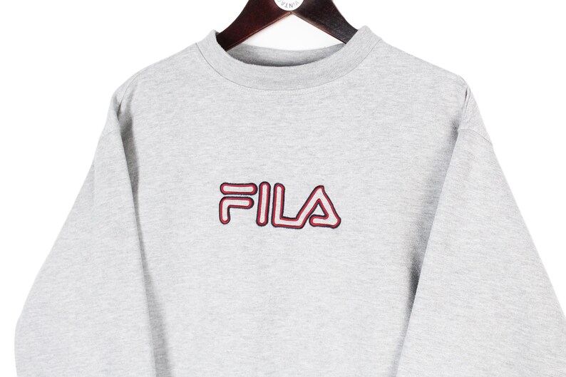 Puede incluir: Sudadera gris claro con el nombre de la marca "FILA" bordado en rojo y rojo oscuro. La sudadera est&aacute; colgada de una percha de madera. La prenda tiene cuello redondo y mangas largas.