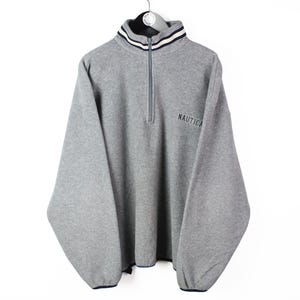 Könnte beinhalten: Ein grauer Fleece-Pullover mit Viertelreißverschluss, langen Ärmeln und einem hohen Kragen mit schwarz-weiß gestreiftem Besatz. Das Wort NAUTICA ist in Schwarz auf der linken Brust gestickt. Das Kleidungsstück hat elastische Ärmelbündchen.