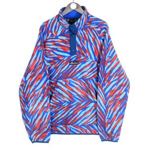 Pode incluir: Um pulôver de lã polar com padrão azul, vermelho e azul claro, com gola alta e carcela abotoada. A parte superior de manga comprida tem um bolso canguru e um pequeno patch da marca.