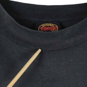 May include: Close-up of a dark gray crew neck sweatshirt. The collar is ribbed and the tag reads "Guinness" in a red and gold design. A wooden stick points towards the collar.