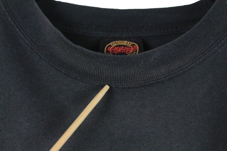 May include: Close-up of a dark gray crew neck sweatshirt. The collar is ribbed and the tag reads "Guinness" in a red and gold design. A wooden stick points towards the collar.