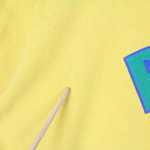 May include: Close-up of a yellow t-shirt with a large, stylized letter "B" in green with a blue outline and blue fill. The fabric has a slightly textured appearance.