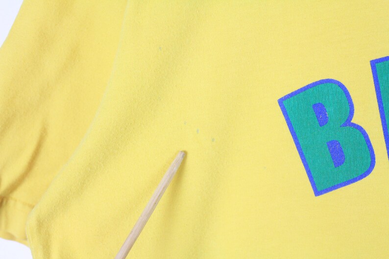 May include: Close-up of a yellow t-shirt with a large, stylized letter "B" in green with a blue outline and blue fill. The fabric has a slightly textured appearance.