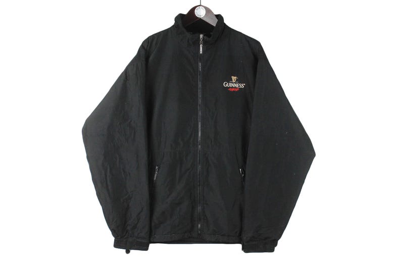K&ouml;nnte beinhalten: Schwarze Rei&szlig;verschlussjacke mit einem Guinness-Logo auf der linken Brust. Das Logo ist in Gold, Rot und Wei&szlig;.