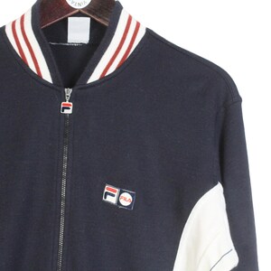 May include: Navy blue zip-up jacket with white and red striped collar and cuffs. Features a small Fila logo patch on the chest. The jacket has white panels on the sides and a full-length zipper.