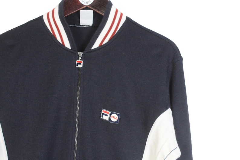 May include: Navy blue zip-up jacket with white and red striped collar and cuffs. Features a small Fila logo patch on the chest. The jacket has white panels on the sides and a full-length zipper.