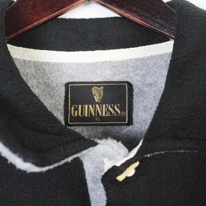 May include: Black fleece jacket with a white trim and a button closure. The jacket features a Guinness label with a harp logo and the text "GUINNESS XL". The collar is black.