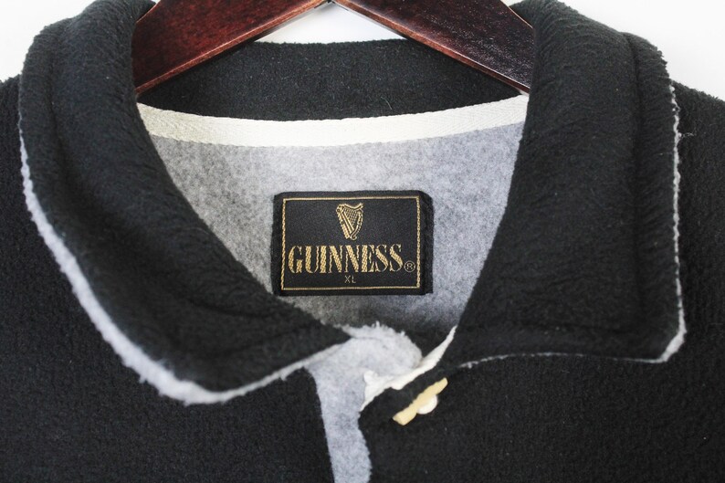 May include: Black fleece jacket with a white trim and a button closure. The jacket features a Guinness label with a harp logo and the text "GUINNESS XL". The collar is black.