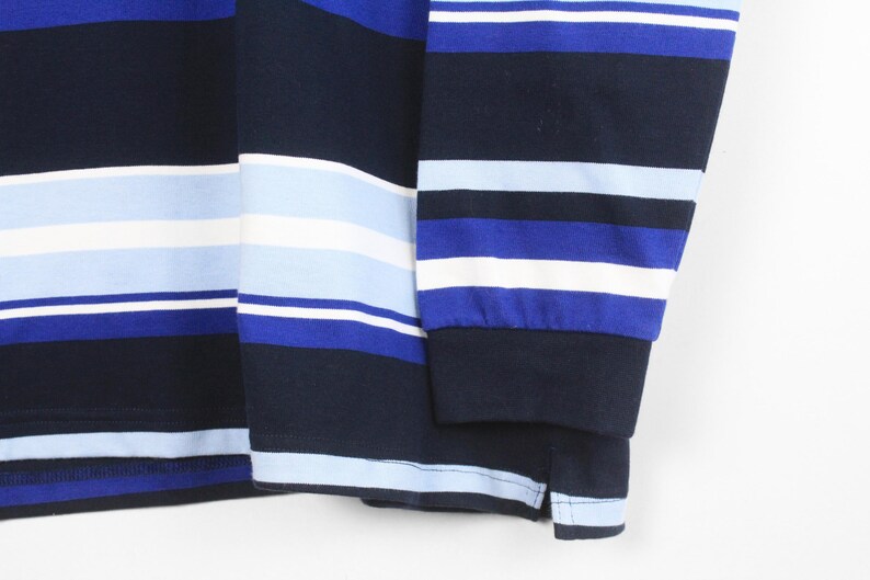 May include: Close-up of a long-sleeved shirt with horizontal stripes in navy blue, white, and various shades of blue. The sleeve cuff is visible, and the fabric appears to be a soft knit.