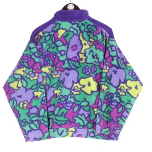 Puede incluir: Una chaqueta de forro polar de manga larga con un vibrante estampado floral en tonos morado, amarillo y verde sobre un fondo verde claro. La chaqueta tiene un cuello y paneles en los hombros de color morado s&oacute;lido, cremallera frontal y pu&ntilde;os acanalados.