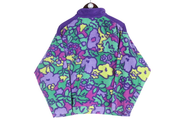 Puede incluir: Una chaqueta de forro polar de manga larga con un vibrante estampado floral en tonos morado, amarillo y verde sobre un fondo verde claro. La chaqueta tiene un cuello y paneles en los hombros de color morado s&oacute;lido, cremallera frontal y pu&ntilde;os acanalados.