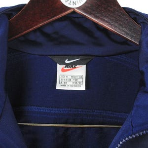 May include: Close-up of a navy blue Nike track jacket with a zipper and a collar. The Nike logo is visible on a tag, along with size information. The jacket is hanging on a wooden hanger.