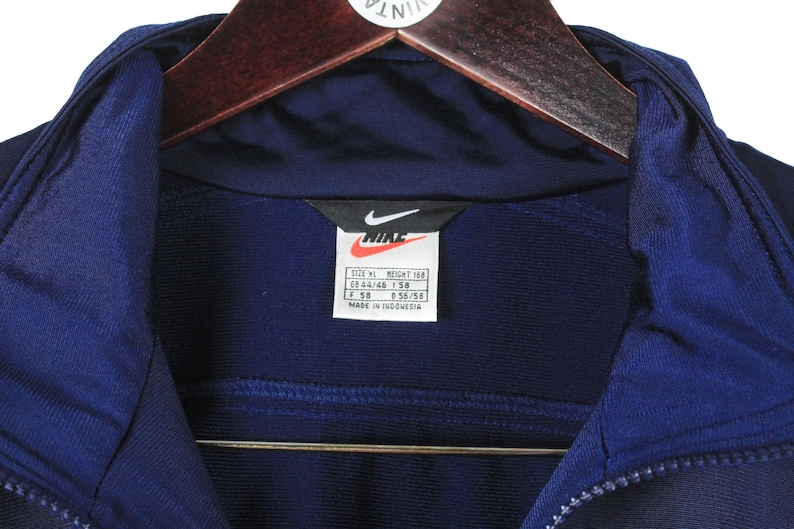 May include: Close-up of a navy blue Nike track jacket with a zipper and a collar. The Nike logo is visible on a tag, along with size information. The jacket is hanging on a wooden hanger.