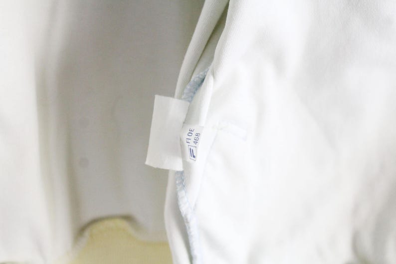May include: A white fabric garment interior featuring a small, rectangular white care label. The label displays the text FI DE 468 in black ink, positioned above a small blue and white logo icon. The fabric appears smooth and clean.