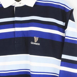 May include: A long-sleeved rugby shirt with navy, white, and blue horizontal stripes. It has a white collar and the Guinness logo embroidered on the chest. The shirt is a classic style.
