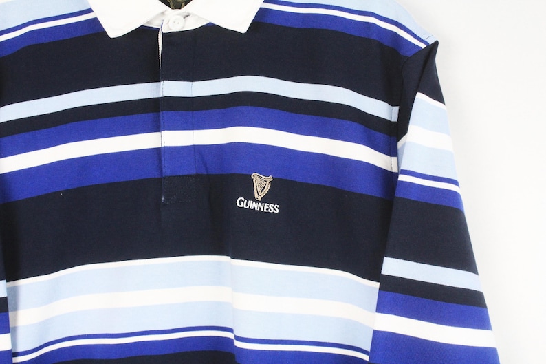 May include: A long-sleeved rugby shirt with navy, white, and blue horizontal stripes. It has a white collar and the Guinness logo embroidered on the chest. The shirt is a classic style.