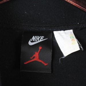 Może przedstawiać: Czarna bluza Nike z białym logo Nike i czerwonym logo Jumpman na czarnej metce.