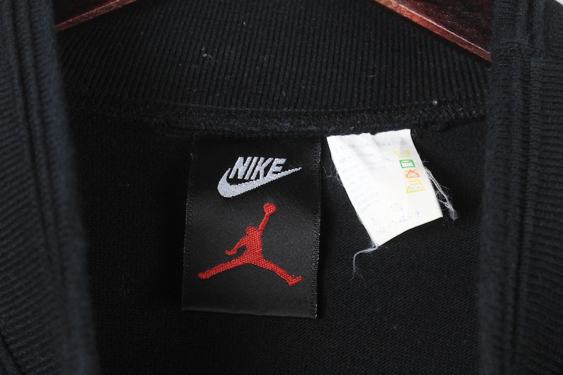 Może przedstawiać: Czarna bluza Nike z białym logo Nike i czerwonym logo Jumpman na czarnej metce.