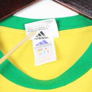 May include: A yellow t-shirt with a green collar and trim, hanging on a wooden hanger. The Adidas logo is visible on a white tag. The tag also includes text and international flags.