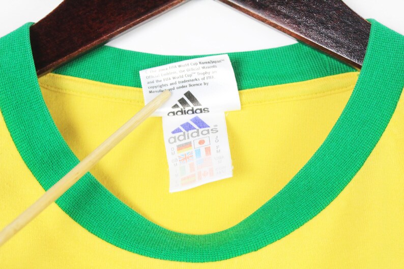 May include: A yellow t-shirt with a green collar and trim, hanging on a wooden hanger. The Adidas logo is visible on a white tag. The tag also includes text and international flags.