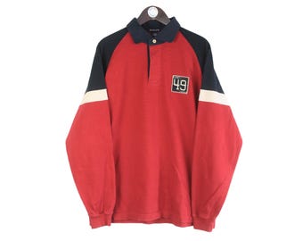 May include: A red rugby shirt with navy and white detailing. The long-sleeved shirt has a navy collar and sleeves, with a white stripe on the upper arm. A patch with the number "49" is on the chest.