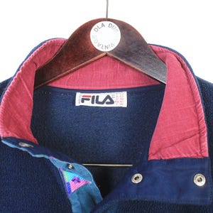 K&ouml;nnte beinhalten: Marineblaue Fleecejacke mit bordeauxfarbenem Kragen und Akzenten. Die Jacke hat einen Knopfverschluss und ein teal- und lilafarbenes Musterdetail. Das Fila-Logo ist auf dem Etikett sichtbar. Die Jacke h&auml;ngt an einem Holzkleiderb&uuml;gel.